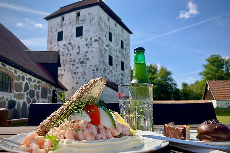 Bild på en stor räksmörgås, en drick och två kakor framför porttornet på Hovdala
