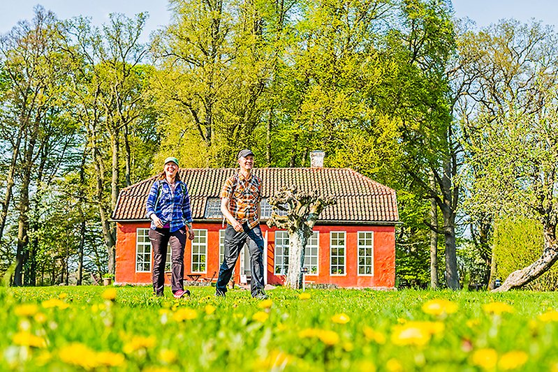 Två vandringsklädda personer går genom slottsparken med det röda orangeriet i bakgrunden.