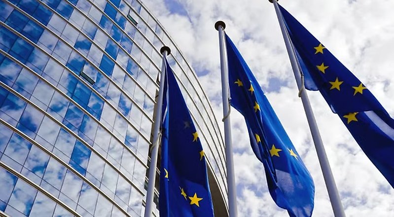 Bild på EU-parlamentet med tre EU-flaggor som vajar i vinden. 